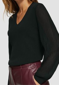 Kobieta w czarnym swetrze z dekoltem w serek, z przezroczystymi, pionowymi paskami na długich rękawach oraz w wysokich, burgundowych skórzanych spodniach, z ręką w kieszeni.