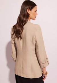 Beżowa lniana marynarka z podwiniętymi mankietami i smukłą sylwetką. Posiada delikatnie teksturowany materiał oraz minimalne przeszycia.