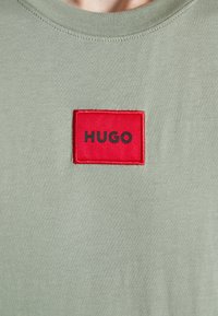 Camiseta de algodón verde oliva con un cuello redondo y un prominente parche rectangular rojo que muestra "HUGO" en letras negras.