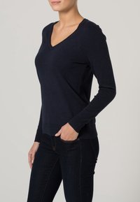 Marineblauer Strickpullover mit V-Ausschnitt und langen Ärmeln. Gerippte Bündchen und Saum. Über dunklen Jeans getragen, was eine figurbetonte Silhouette betont.