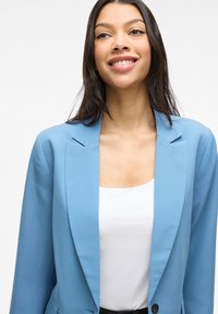 Svetlomodrý blazer s klopami, s jedným gombíkom na zapínanie a hladkou textúrou, nosený cez biely top, v úzkom strihu.