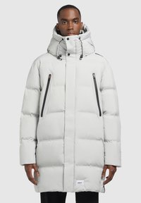 Férfi világosszürke hosszú steppelt kabátban, magas gallérral, kapucnival és két függőleges, fekete cipzáros zsebből álló zsebeivel, szürke háttér előtt áll.