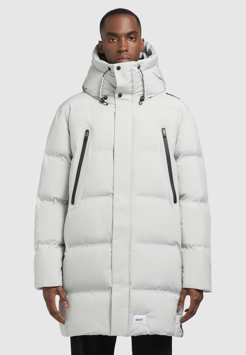 Férfi világosszürke hosszú steppelt kabátban, magas gallérral, kapucnival és két függőleges, fekete cipzáros zsebből álló zsebeivel, szürke háttér előtt áll.