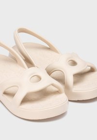 Sandales en caoutchouc beige avec un semelle intérieure texturée, bout ouvert et deux brides comportant des découpes circulaires pour la respirabilité et le soutien.