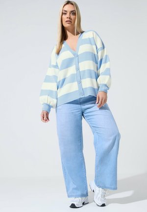 Femme portant un cardigan rayé bleu et blanc, un jean large bleu clair et des baskets blanches, debout devant un fond clair uni.