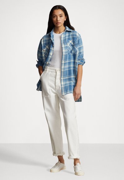 Camicia a scacchi blu con due tasche sul petto, indossata sopra una t-shirt bianca e abbinata a pantaloni bianchi con risvolto e sneakers bianche. Texture liscia.