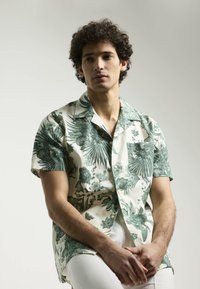 Camisa de manga corta y botones, con un patrón de flores y hojas verdes sobre un fondo beige, y un bolsillo en el lado izquierdo del pecho.