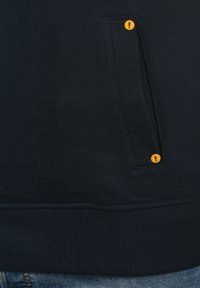 Sudadera azul marino con un dobladillo acanalado horizontal, tejido texturizado y un pequeño botón amarillo en un bolsillo lateral.
