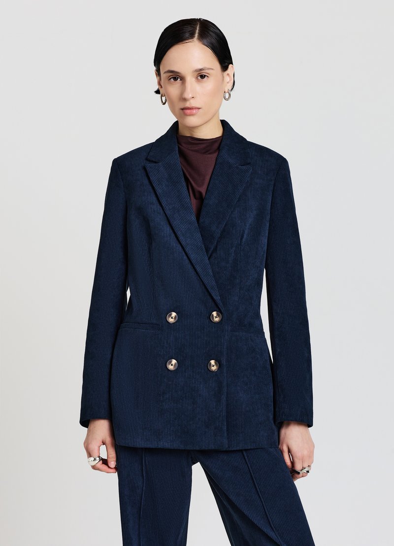 Blazer doppiopetto in velluto a coste blu navy con revers, caratterizzato da bottoni dorati e una silhouette strutturata, abbinato a pantaloni coordinati.