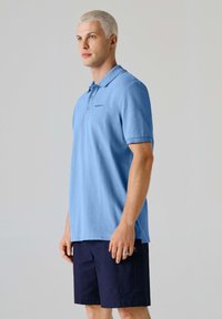 Junger Mann mit kurzen blonden Haaren, der ein hellblaues Poloshirt und dunkelblaue Shorts trägt, vor einem schlichten grauen Hintergrund stehend.