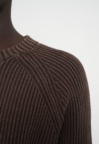 Καφέ ριγωτό πουλόβερ με υφασμένη πλέξη, που διαθέτει στρογγυλή λαιμόκοψη και διαγώνια λεπτομέρεια ραφής στον ώμο. Κοντινή λήψη.