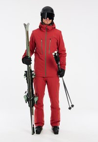 Skiausrüstung in leuchtendem Rot, bestehend aus einer wasserdichten Jacke und Hose. Dazu gehören Skistöcke und Skier mit grünen Akzenten. Schwarzer Helm und Goggles.