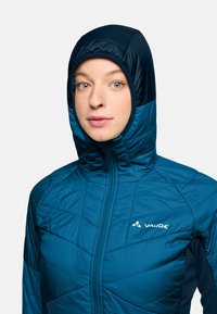 Lichte blauwe geïsoleerde jas met een capuchon, voorzien van een gevoerd patroon, een volledige rits aan de voorkant en het "VAUDE" logo op de borst.