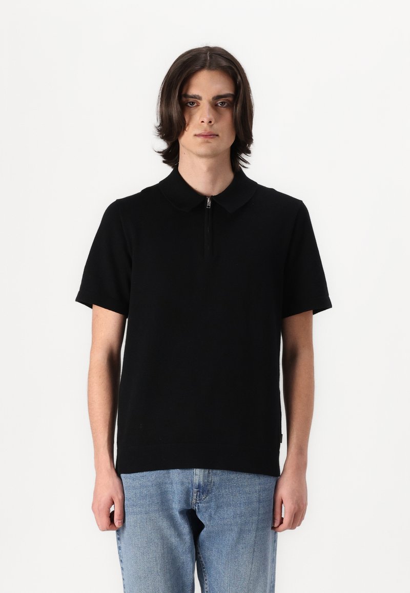 Svart kortermet polo-skjorte med glidelås i kragen. Strikket stoff, avslappet passform, kombinert med lyseblå denimjeans. Enkel design, uten mønstre.