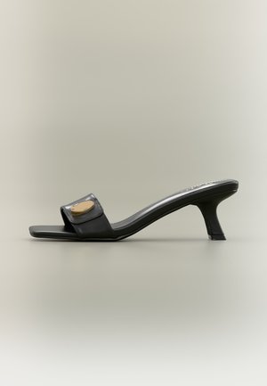 Sort mule-sandal med åben tå, mellemhøj buet hæl og gyldent ovalt pynt på den brede frontrem, vist på en neutral baggrund.