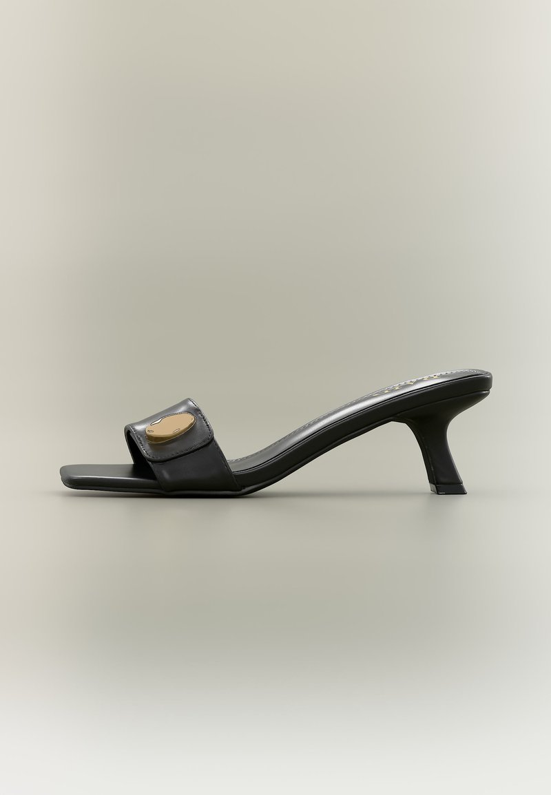 Mule sandale noire à bout ouvert avec talon incurvé de hauteur moyenne et ornement ovale doré sur la large bride avant, posée sur un fond neutre.