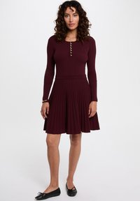 Vestido de canalé burdeos con mangas largas, tapeta de botones, cuerpo ajustado y falda plisada. Llevado con bailarinas negras.