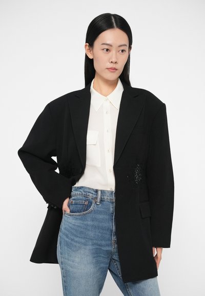 Černé oversized sako s texturovanými detaily, nošené přes bílou košili na knoflíky. Světle modré džínové kalhoty doplňují outfit.