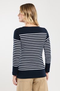 Frau, die nach links schaut, trägt ein navyblaues und weißes horizontal gestreiftes Langarmshirt und beige Hosen vor einem schlichten Hintergrund.