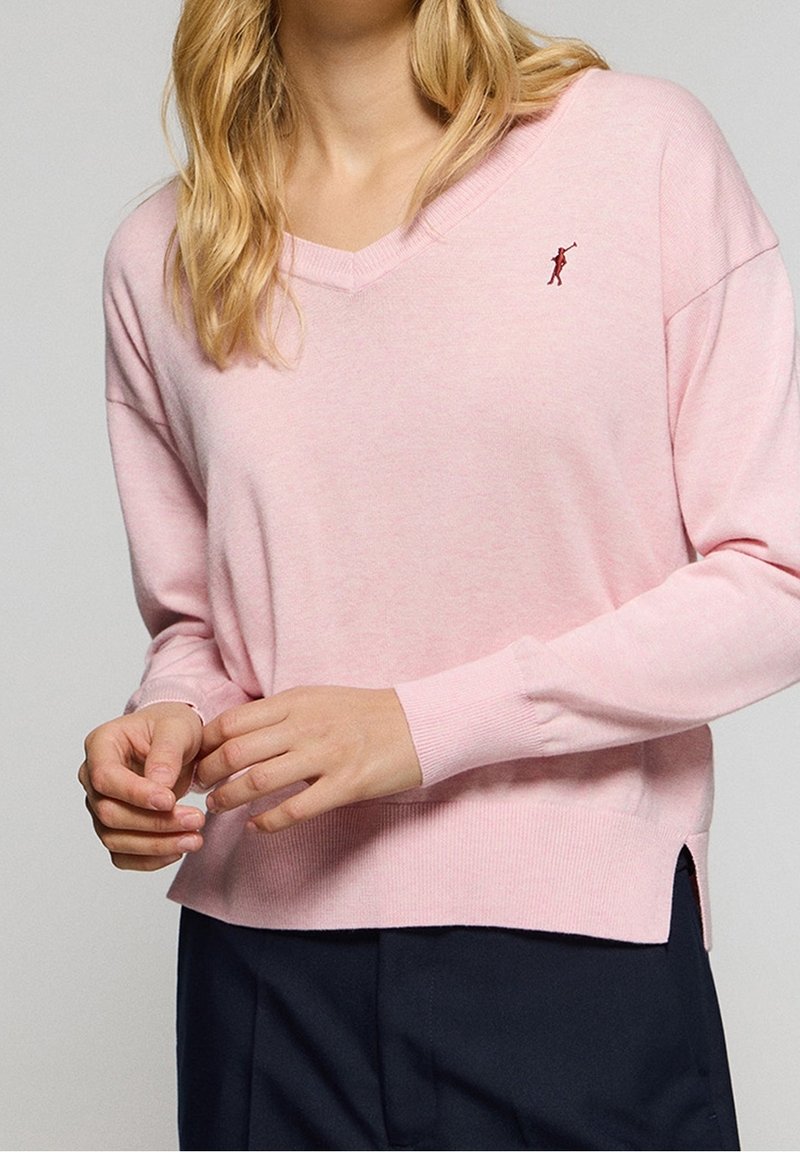 Light pink v-neck sweater with long sleeves, ribbed cuffs, side slits, and a small embroidered detail on the chest. Soft, textured material.
