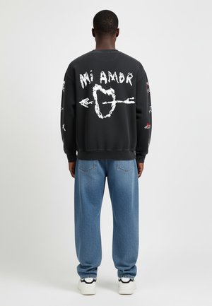 Sweatshirt noir avec le texte graphique blanc "Mi Amor" et un cœur avec une flèche, associé à un pantalon en denim bleu clair et des sneakers blanches.