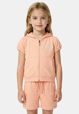 Jeune fille portant un sweat à capuche zippé à manches courtes couleur pêche et un short assorti avec cordon de serrage et poches latérales, debout devant un fond blanc.