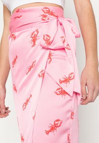 Jupe portefeuille rose en tissu lisse, avec un imprimé homard rouge. Comprend un détail noué à la taille et un design fluide.