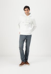 Weißer Hoodie mit grafischem Frontdruck, Kordelzugkapuze und gerippten Bündchen; kombiniert mit blauen Jeans und braunen Sneakern.