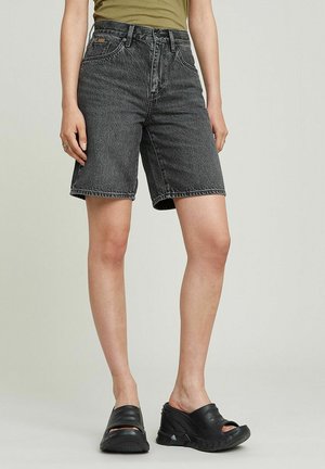 Jeansshorts in dunkler Waschung mit lockerem Schnitt, mittelhohem Bund und sichtbaren Nähdetails. Kombiniert mit schwarzen Slippern.