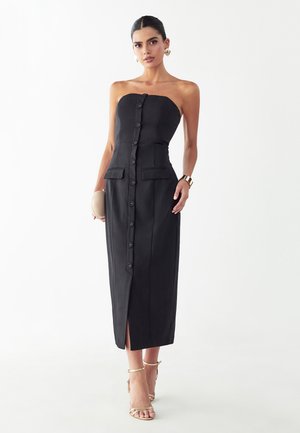 Frau trägt ein trägerloses, schwarzes Midikleid mit vorderen Knöpfen, Pattentaschen, goldenen Ohrringen, Armband, hält eine beige Clutch und trägt goldene Sandaletten mit hohen Absätzen.