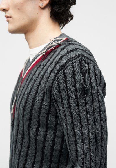 Jeune personne portant un pull gris à torsades avec des rayures rouges et des fils lâches le long des bords des épaules et du col, regardant vers la gauche.