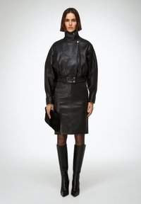 Mujer de pie con chaqueta negra de cuero de cuello alto y cintura elástica, falda a juego hasta la rodilla, medias negras transparentes, botas negras hasta la rodilla, sosteniendo un bolso negro de mano.