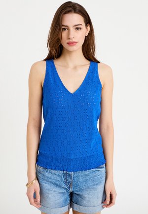 Ung kvinde iført en blå ærmeløs tekstureret top med V-hals og opkørte lyse denimshorts, stående mod en ensartet hvid baggrund.