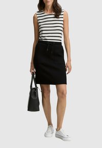 Femme portant un haut sans manches blanc et noir à rayures, une jupe noire à cordon, des baskets blanches, tenant un sac à main noir sur le côté.