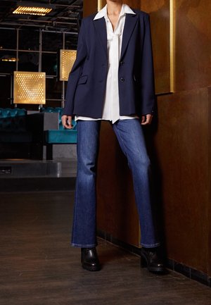 Person standing indoors wearing a navy blazer, white collared shirt, blue jeans, and black platform boots against a bronze wall background.