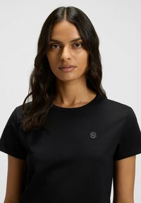 Schwarzes Baumwoll-T-Shirt mit kurzen Ärmeln und rundem Ausschnitt. Verfügt über ein dezentes gesticktes Logo auf der linken Brustseite. Glatte Textur.