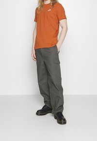 Oranžové bavlněné tričko s bílým logem, ve spojení s tmavě zelenými cargo kalhotami se bočními kapsami a černými botami se žlutými akcenty.