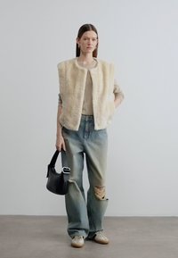 Γιλέκο από συνθετική γούνα σε κρεμ, μπεζ πλεκτό τοπ, φθαρμένο τζιν ανοιχτού χρώματος, μαύρη τσάντα χειρός και μπεζ αθλητικά με χοντρή σόλα.