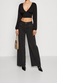 Svart långärmad crop top med V-ringning, kombinerad med vida svarta jeans och svarta högklackade skor. Inkluderar en beige handväska.