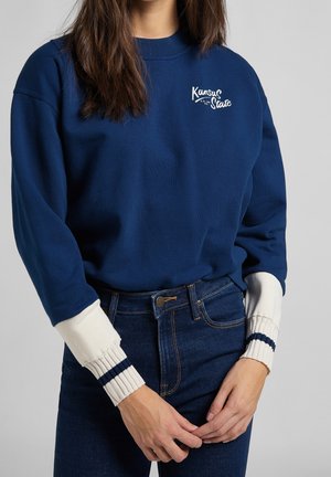 Sudadera azul marino con bordado blanco de "Kansas State", puños de canalé crema en contraste y corte relajado, combinada con jeans de mezclilla oscuros.