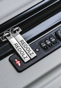 Twee metalen ritssluitingen met het label "REDOLZ" op een zilverkleurige, gestructureerde koffer bij een cijferslot met drie cijfers.