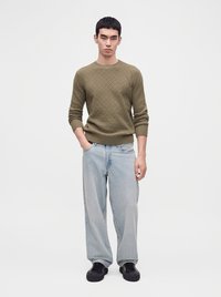 Giovane uomo in piedi che indossa un maglione verde oliva con trama, jeans larghi azzurri chiari e sneakers nere su uno sfondo bianco uniforme.