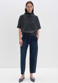 Donna in piedi che indossa un top corto grigio scuro a maniche corte, jeans aderenti blu scuro a vita alta e ballerine grigie con punta, su uno sfondo bianco.