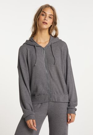 Junge Frau trägt einen grauen Waffelstrick-Zip-Hoodie und passende Hose und steht vor einem schlichten weißen Hintergrund.