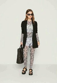 Blazer preto sobre uma blusa clara e estampada com motivos de borboleta, leggings a condizer, bolsa preta e sandálias chunky. Detalhes de tecido texturizado.