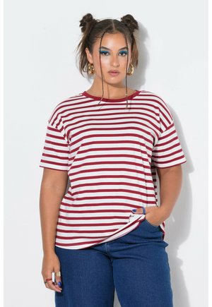 Frau mit geflochtenen Space Buns, die goldene Creolen trägt, ein rot-weiß gestreiftes T-Shirt, blaue Jeans und blaues Augen-Make-up trägt und mit einer Hand in der Tasche posiert.
