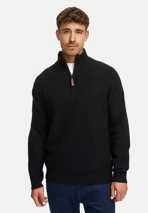 Junger Mann, der einen schwarzen Strickpullover mit halbem Reißverschluss und dunkelblauen Jeans trägt, steht vor einem schlichten weißen Hintergrund.