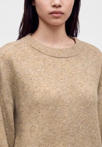 Femme portant un pull en laine beige à col rond, aux longs cheveux foncés, montrée des épaules jusqu'à la bouche sur un fond uni.