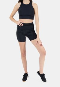 Mustat urheilutoppi korkealla kauluksella ja elastisella vyötäröllä, yhdistettynä mustiin korkeavyötäröisiin shortseihin. Mallilla mustat urheilunikenkä.