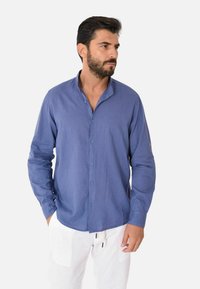 Camicia azzurra con bottoni in tessuto leggero, caratterizzata da un collo semi-alto, maniche lunghe e una vestibilità rilassata. Indossata con pantaloni bianchi.